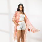 A woman modeling a lightweight peach and white patternedkaftan cover-up over a white crop top and white shorts, with sunglasses on her head, set against a bright white studio background.