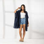 A woman modeling a dark navy blue open-front Kaftan with a lace trim over a white crop top and denim shorts, standing against a white wall with soft window shadows.