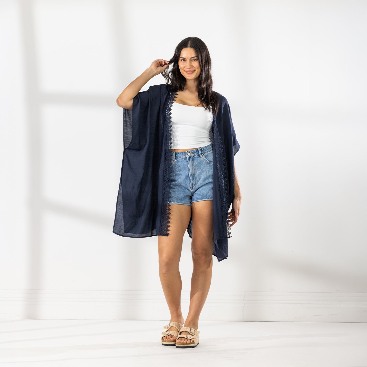 A woman modeling a dark navy blue open-front Kaftan with a lace trim over a white crop top and denim shorts, standing against a white wall with soft window shadows.