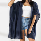 A woman modeling a dark navy blue open-front Kaftan with a lace trim over a white crop top and denim shorts, standing against a white wall with soft window shadows.
