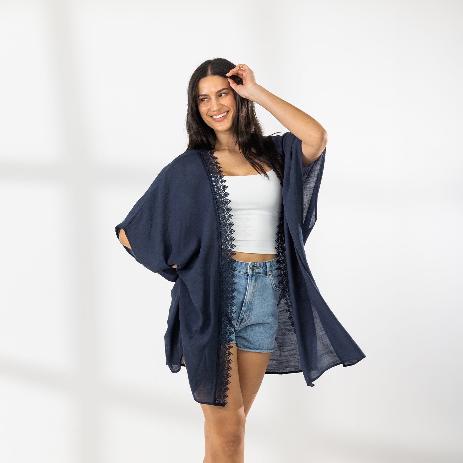 A woman modeling a dark navy blue open-front Kaftan with a lace trim over a white crop top and denim shorts, standing against a white wall with soft window shadows.