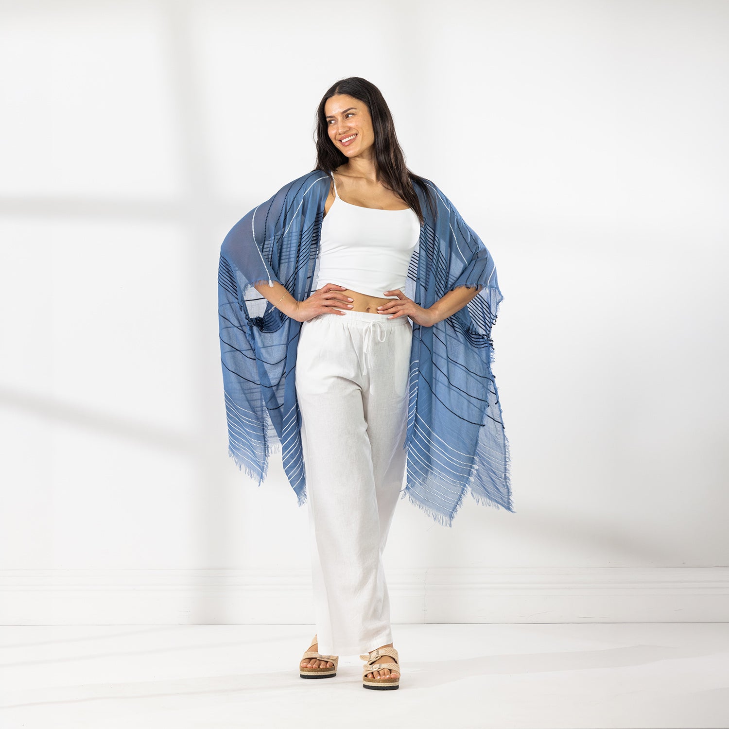 A woman modeling a Chambray striped fringed Kaftan or wrap over a white crop top and white Pants, standing against a white studio wall.