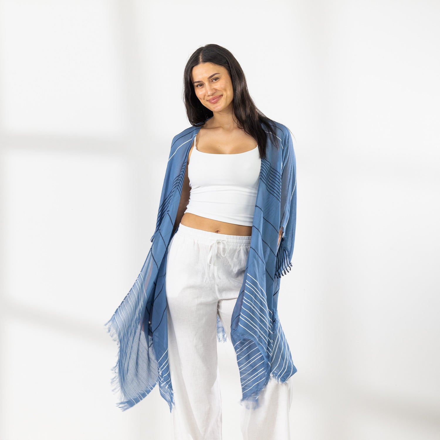 A woman modeling a Chambray striped fringed Kaftan or wrap over a white crop top and white Pants, standing against a white studio wall.