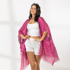 A woman modeling a bright pink and magenta striped fringed Kaftan or wrap over a white crop top and white shorts, standing against a white studio wall.