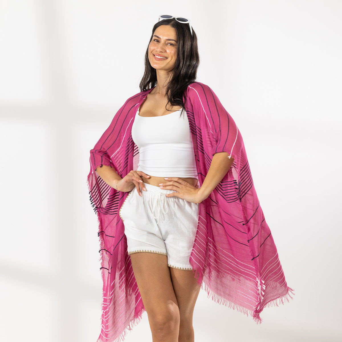A woman modeling a bright pink and magenta striped fringed Kaftan or wrap over a white crop top and white shorts, standing against a white studio wall.