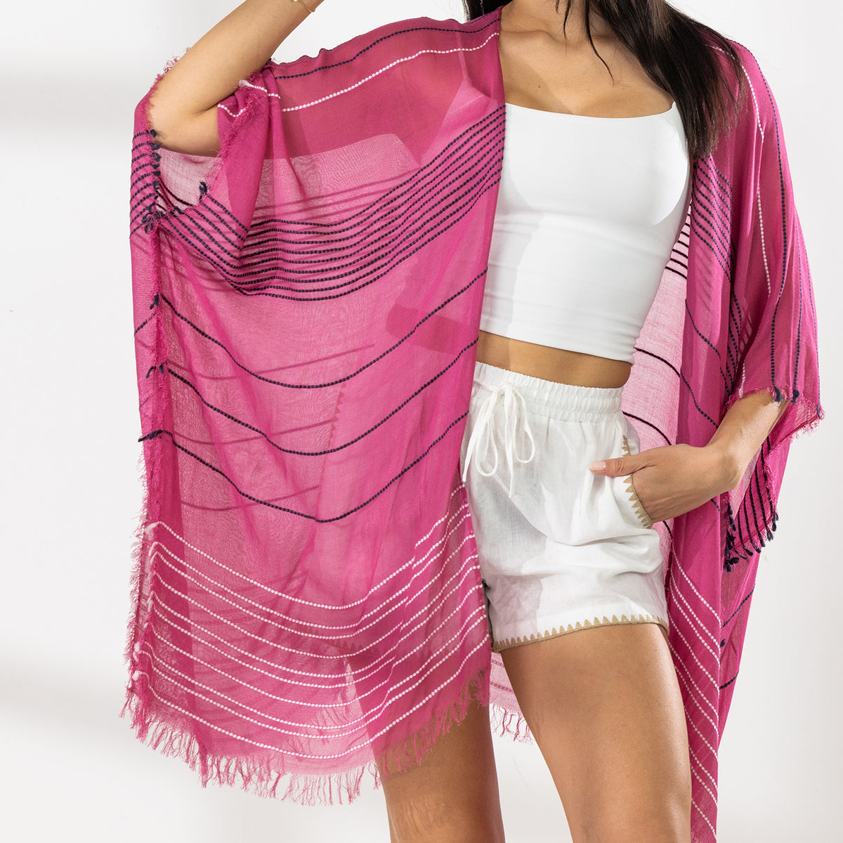 Close Up of A woman modeling a bright pink and magenta striped fringed Kaftan or wrap over a white crop top and white shorts, standing against a white studio wall.