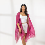 A woman modeling a bright pink and magenta striped fringed Kaftan or wrap over a white crop top and white shorts, standing against a white studio wall.