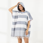 A woman wearing a classic stripe hooded beach poncho with fringed tassels along the hem, photographed against a clean white studio background.