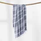 A midnight and white striped beach towel with fringed edges hangs from a thick rope against a clean white background.