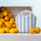 A blue stripe lunch tote with top handles sits on a light wood surface beside a crate of lemons and oranges against a pale blue background.