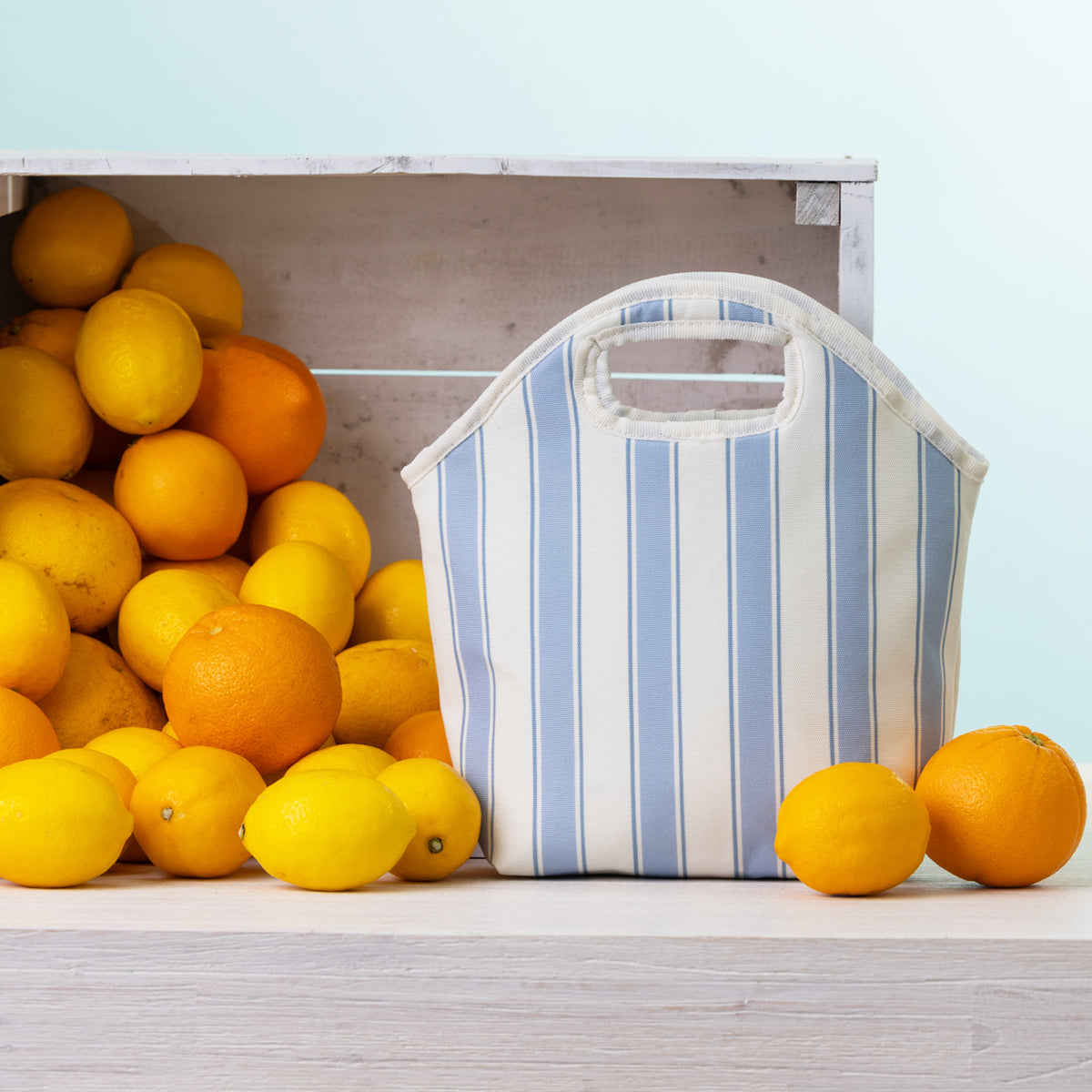 A blue stripe lunch tote with top handles sits on a light wood surface beside a crate of lemons and oranges against a pale blue background.
