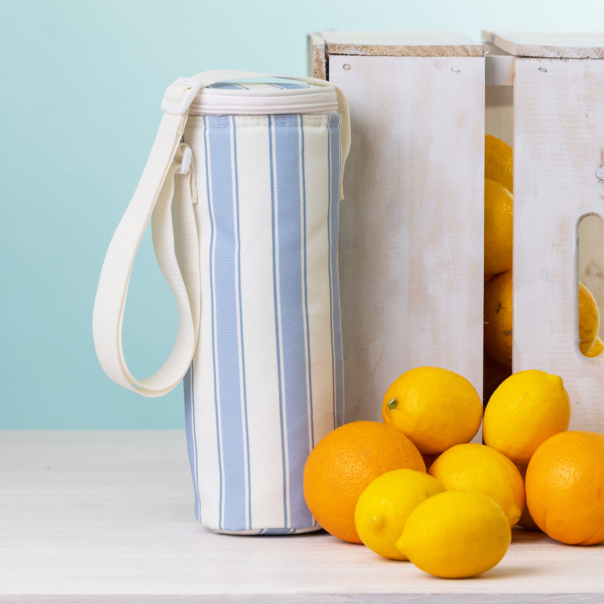 A stripe bottle bag with a white strap stands on a white tabletop beside white wooden crates and bright lemons and oranges against a pale blue background.