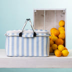 A striped picnic cooler basket sits on a white wooden table next to a white crate filled with oranges, against a light blue background.