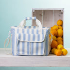 A blue stripe cooler bag sits on a white wooden table next to a white crate filled with oranges, against a light blue background.