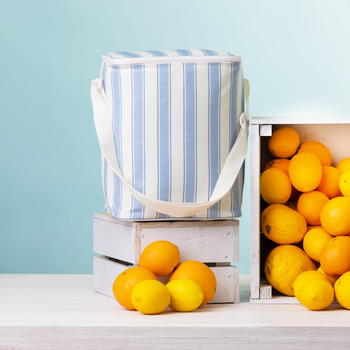 A stripe print duffle bag with a white strap stands on top of a white wooden crate surrounded by bright lemons and oranges against a pale blue background.