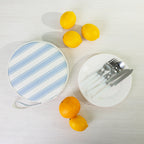 A round fabric picnic bag set with a white plates of silver cutlery styled with citrus fruits on a light wood background.