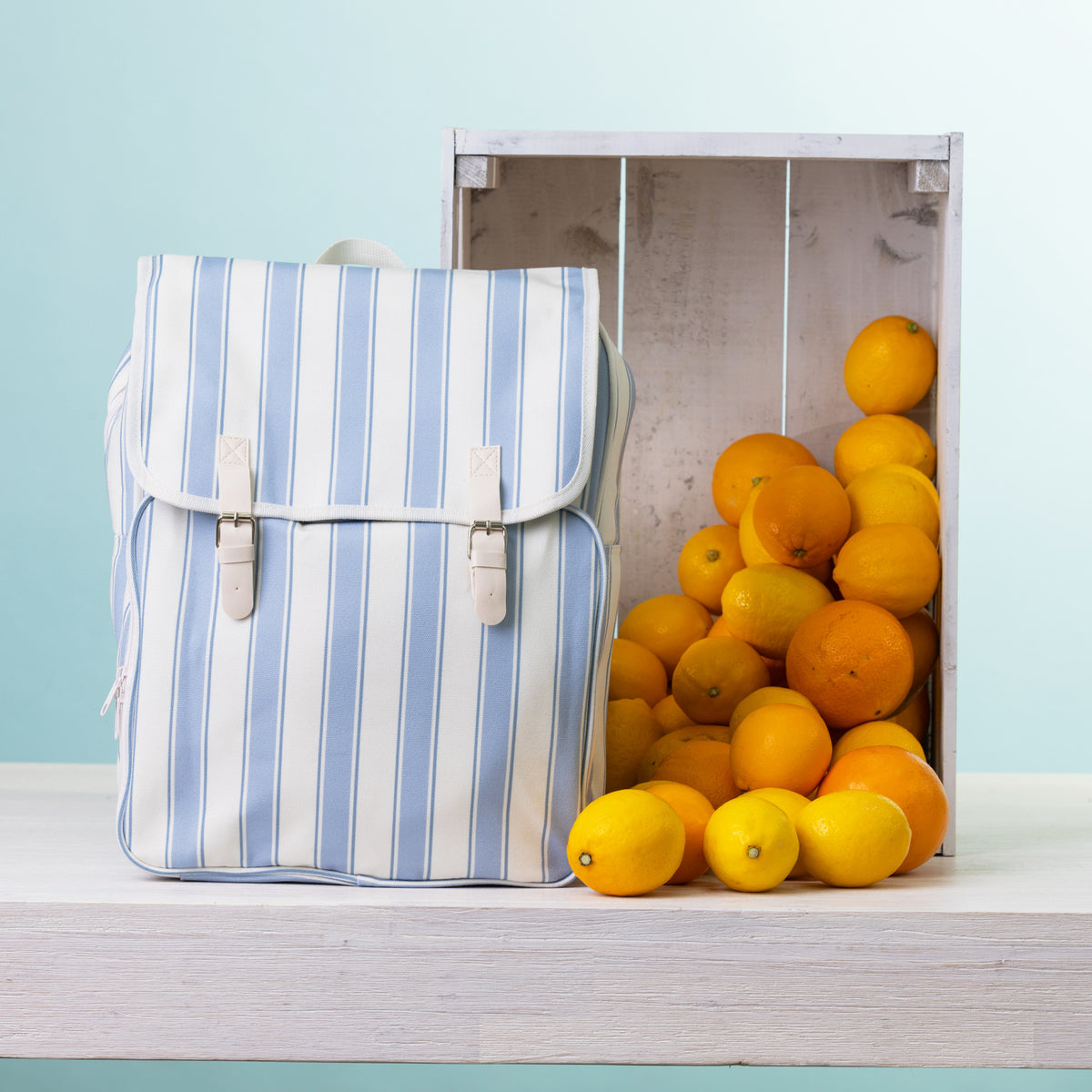 A picnic backpack, styled next to a wooden crate filled with oranges and lemons on a white shelf against a light blue background.