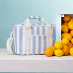 A stripe lunch bag with handles sits on a light wood surface beside a crate of lemons and oranges against a pale blue background.