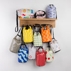 Seaside Supplies Waterproof Beach Bags Range featuring assorted 8L and 5.8L waterproof bags in colourful prints, displayed on a wooden wall shelf with hanging straps.