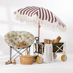 Brown and white striped umbrella with cream fringe trim styled with a beach chair, picnic basket and beach trolley.