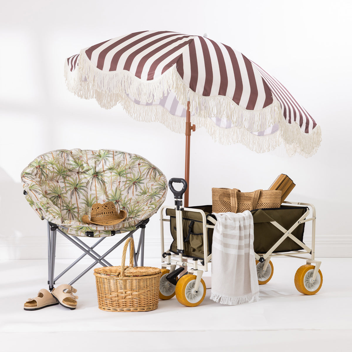 Brown and white striped umbrella with cream fringe trim styled with a beach chair, picnic basket and beach trolley.