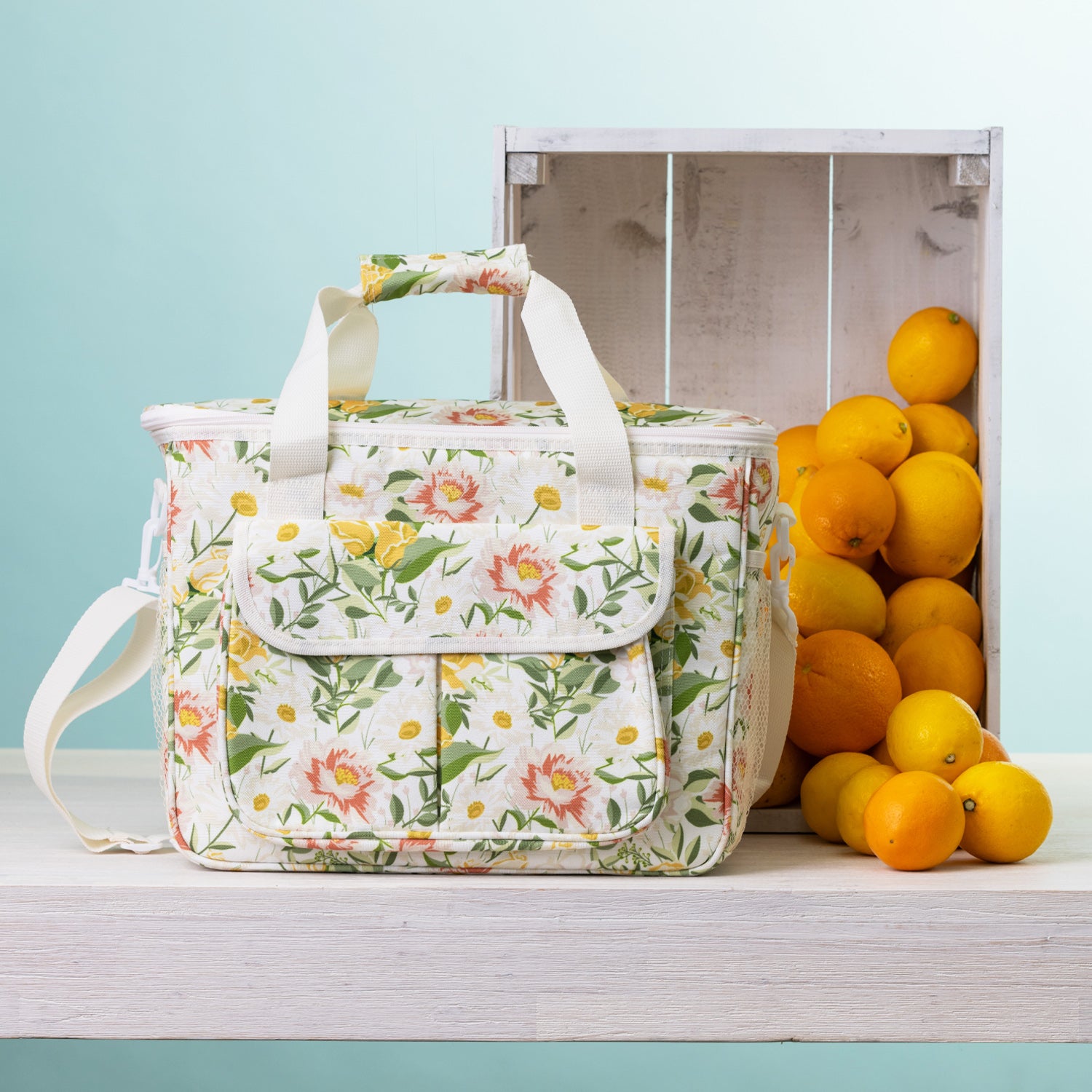 A floral cooler bag sits on a white wooden table next to a white crate filled with oranges, against a light blue background.