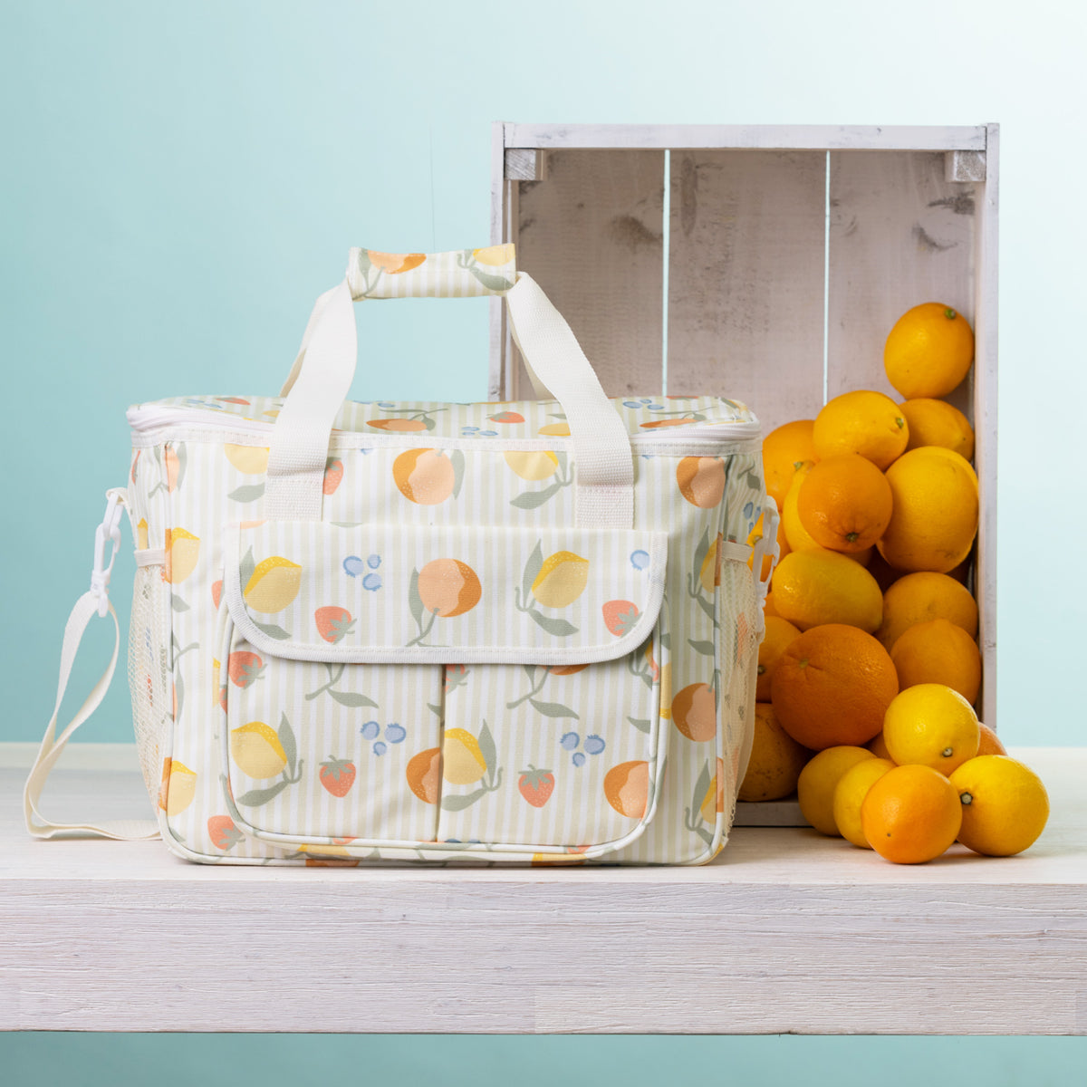 A fruit print cooler bag sits on a white wooden table next to a white crate filled with oranges, against a light blue background.