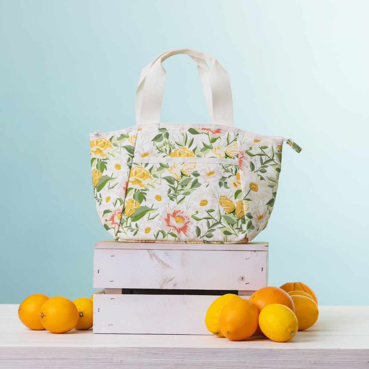 A cooler lunch bag, displayed on a stack of two white wooden crates with oranges and lemons on a light-colored table against a light blue background.