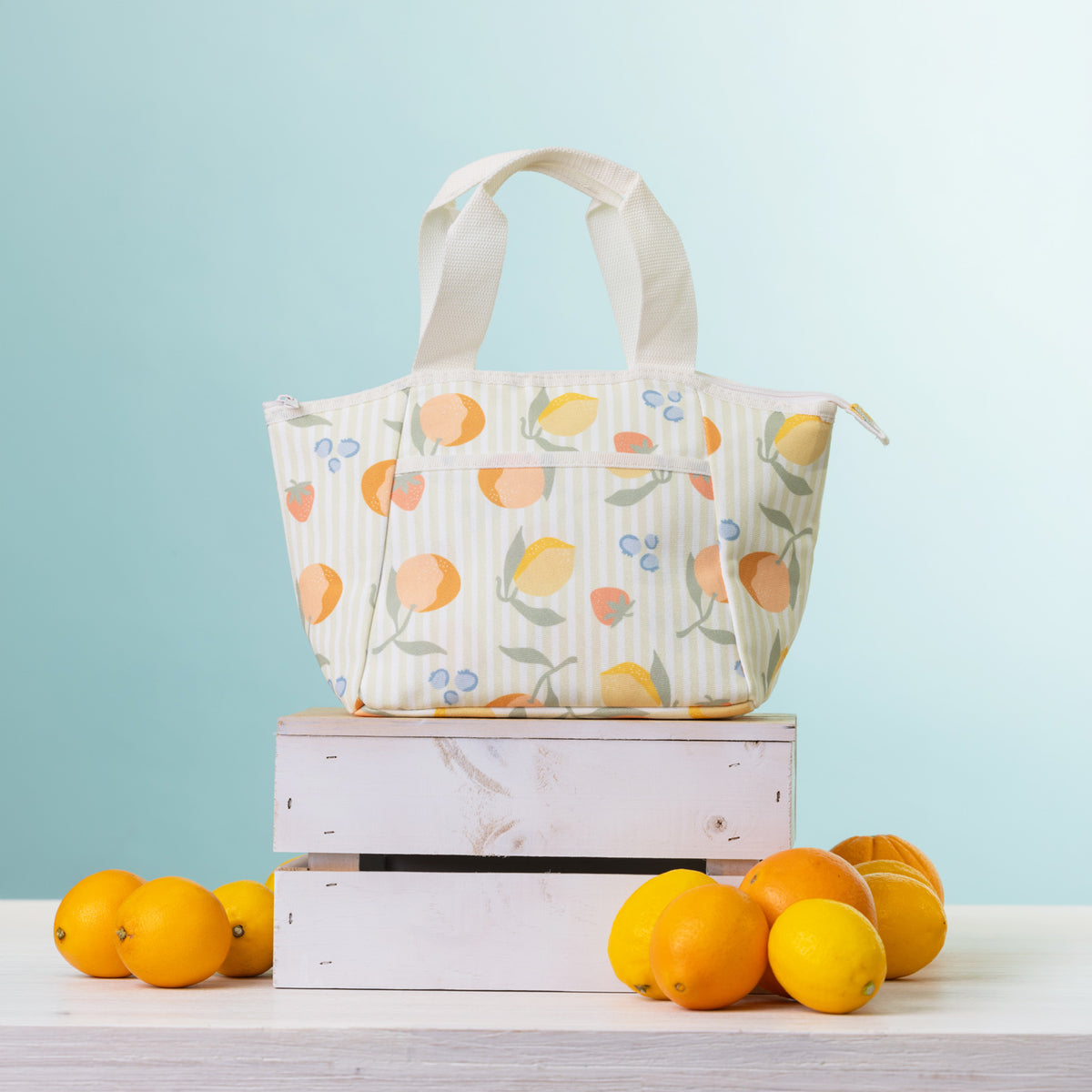 A cooler lunch bag, displayed on a stack of two white wooden crates with oranges and lemons on a light-colored table against a light blue background.