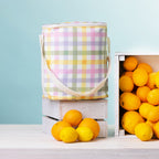 A check print duffle bag with a white strap stands on top of a white wooden crate surrounded by bright lemons and oranges against a pale blue background.