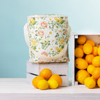 A floral print duffle bag with a white strap stands on top of a white wooden crate surrounded by bright lemons and oranges against a pale blue background.