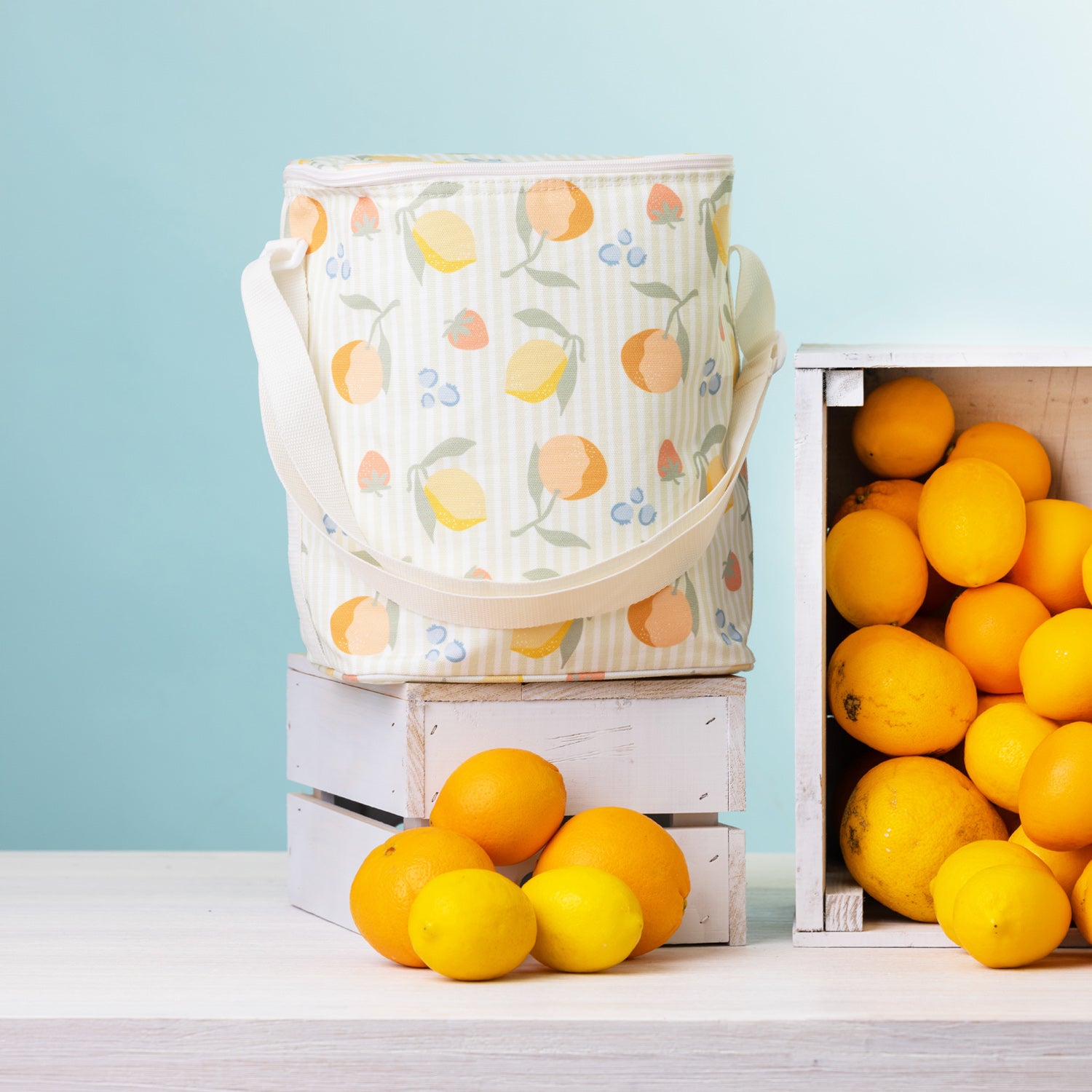 A fruit print duffle bag with a white strap stands on top of a white wooden crate surrounded by bright lemons and oranges against a pale blue background.