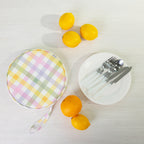 A round fabric picnic bag set with a white plates of silver cutlery styled with citrus fruits on a light wood background.