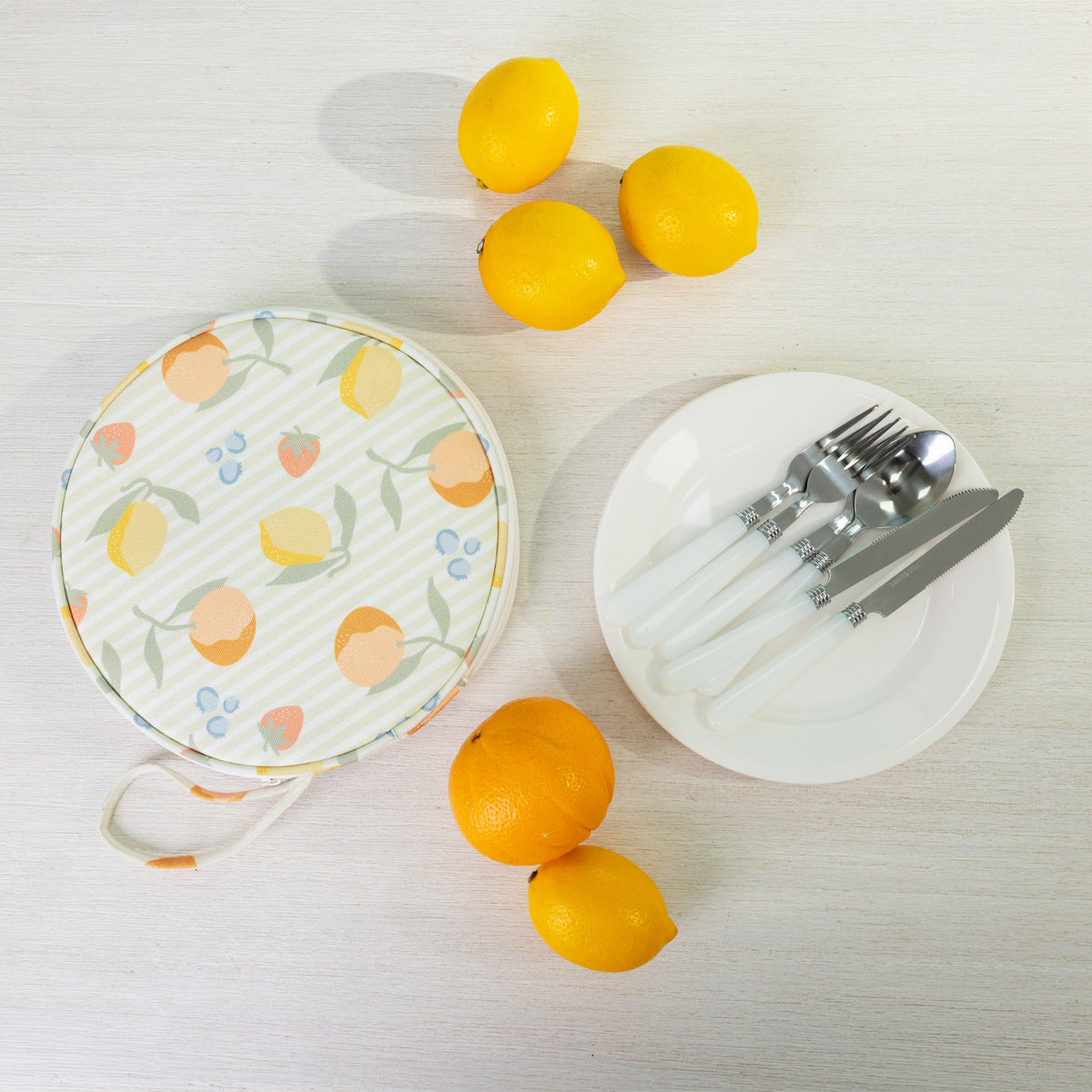 A round fabric picnic bag set with a white plates of silver cutlery styled with citrus fruits on a light wood background.