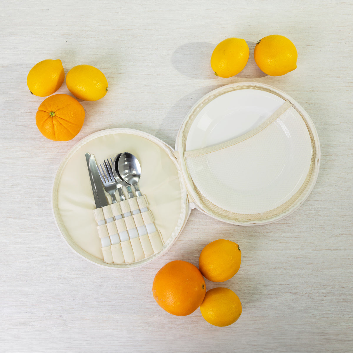 A round fabric picnic bag set with a white plates of silver cutlery styled with citrus fruits on a light wood background.