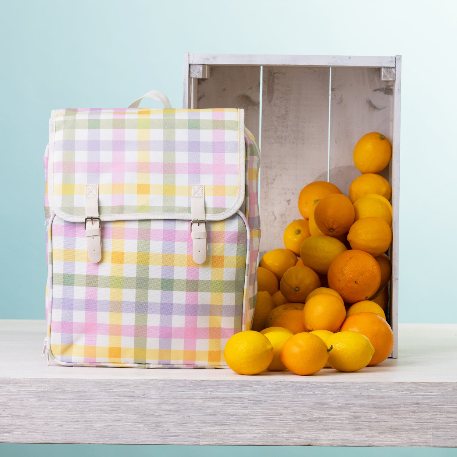 A picnic backpack, styled next to a wooden crate filled with oranges and lemons on a white shelf against a light blue background.