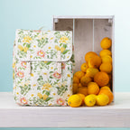 A picnic backpack, styled next to a wooden crate filled with oranges and lemons on a white shelf against a light blue background.