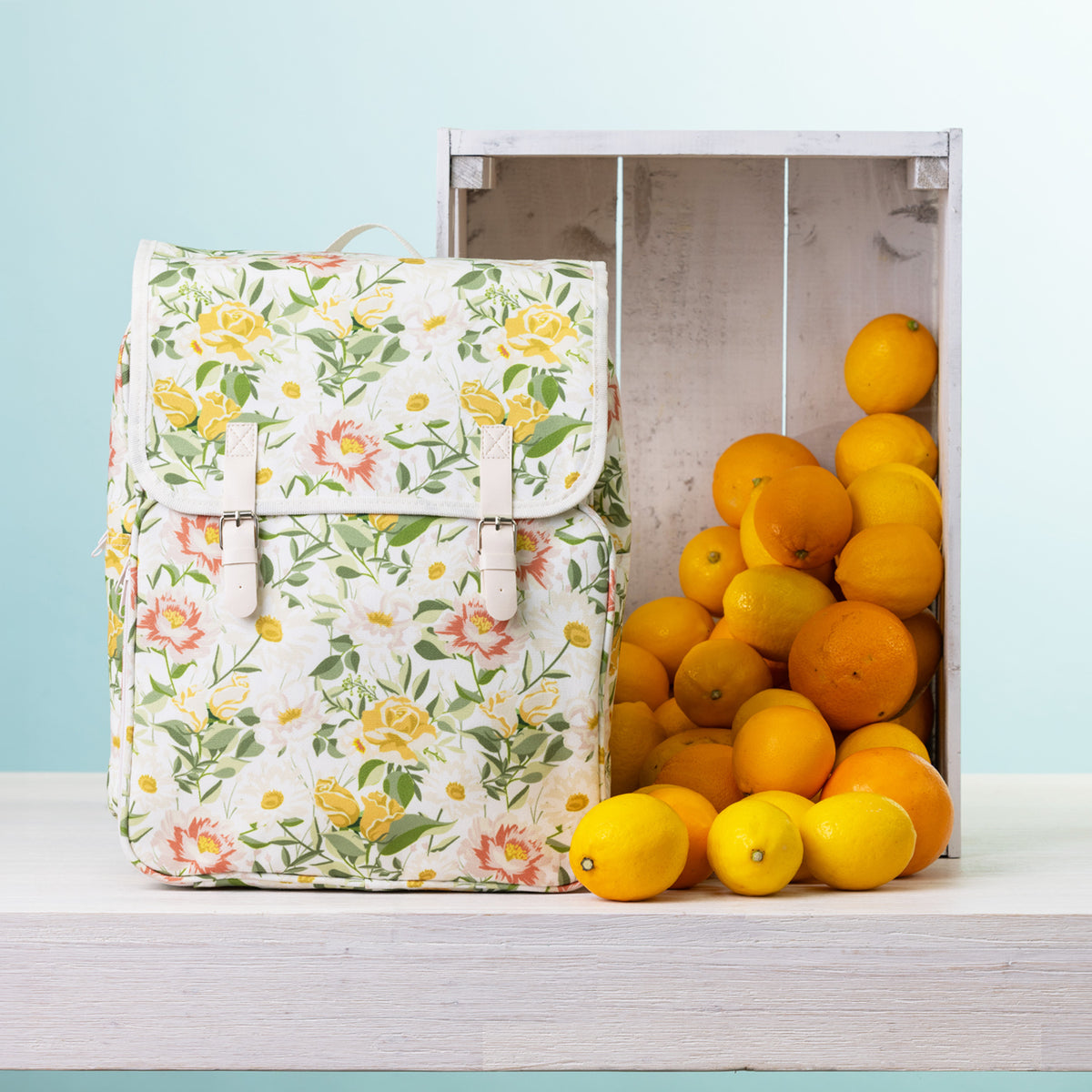 A picnic backpack, styled next to a wooden crate filled with oranges and lemons on a white shelf against a light blue background.