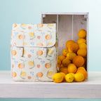 A picnic backpack, styled next to a wooden crate filled with oranges and lemons on a white shelf against a light blue background.