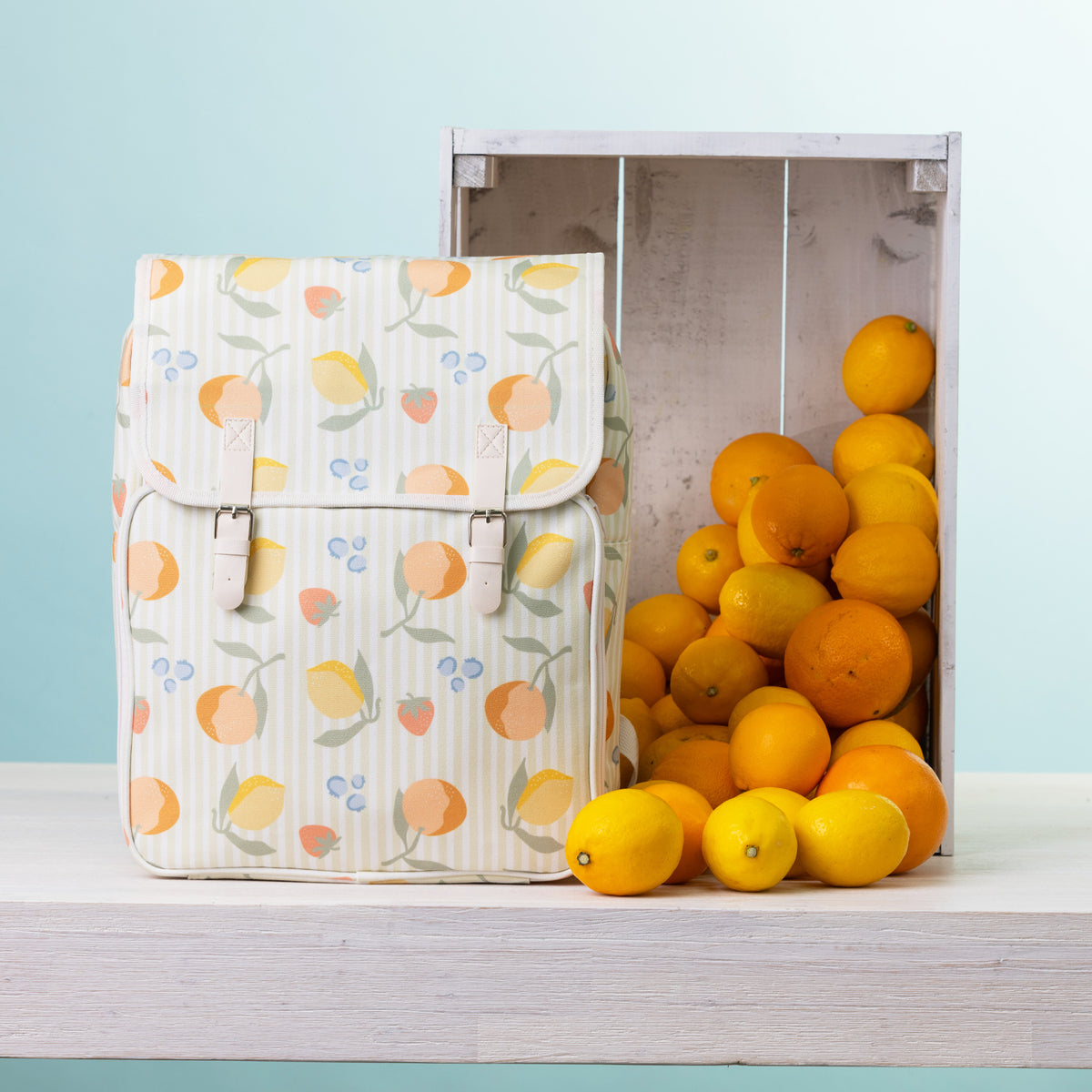 A picnic backpack, styled next to a wooden crate filled with oranges and lemons on a white shelf against a light blue background.