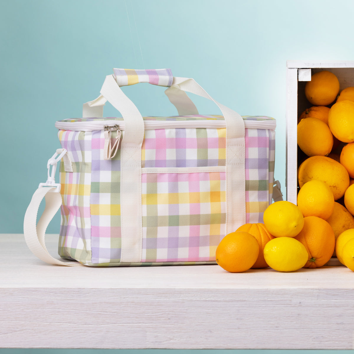 A check lunch bag with handles sits on a light wood surface beside a crate of lemons and oranges against a pale blue background.