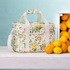 A floral lunch bag with handles sits on a light wood surface beside a crate of lemons and oranges against a pale blue background.