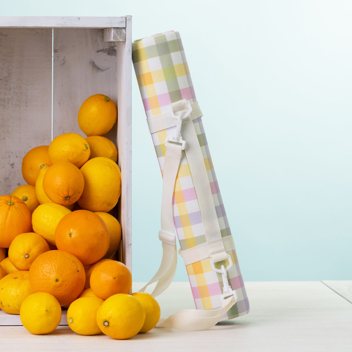 A picnic mat rolled up with a white shoulder strap stands next to a white crate filled with oranges on a light surface against a pale blue background.