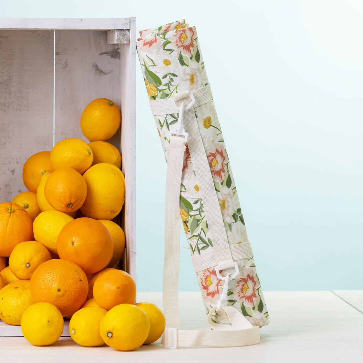 A picnic mat rolled up with a white shoulder strap stands next to a white crate filled with oranges on a light surface against a pale blue background.