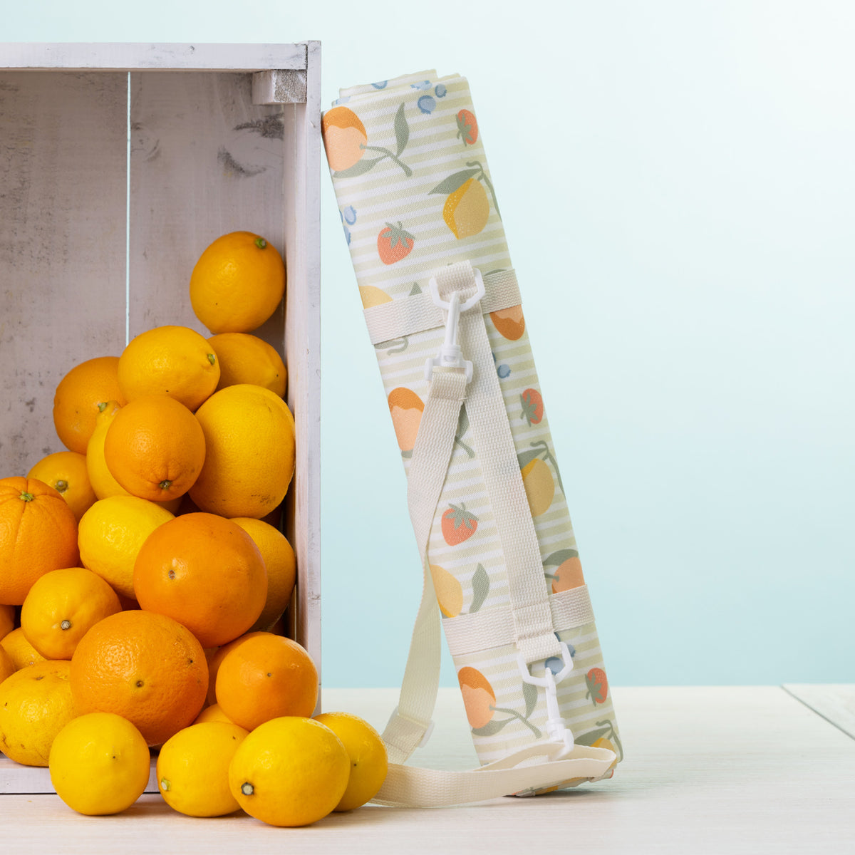 A picnic mat rolled up with a white shoulder strap stands next to a white crate filled with oranges on a light surface against a pale blue background.