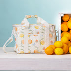 A fruit print lunch bag with handles sits on a light wood surface beside a crate of lemons and oranges against a pale blue background.