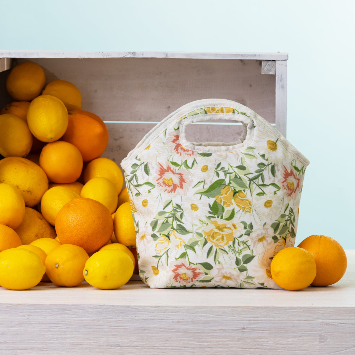 A floral lunch tote with top handles sits on a light wood surface beside a crate of lemons and oranges against a pale blue background.