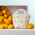 A summer fruits lunch tote with top handles sits on a light wood surface beside a crate of lemons and oranges against a pale blue background.
