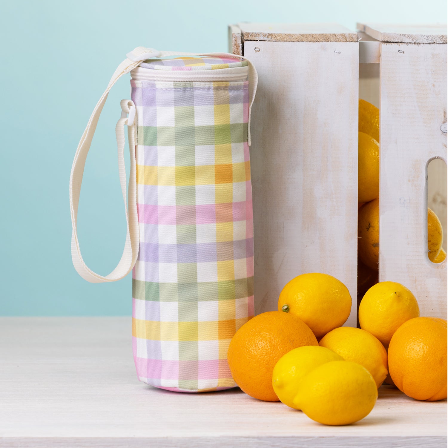A check bottle bag with a white strap stands on a white tabletop beside white wooden crates and bright lemons and oranges against a pale blue background.