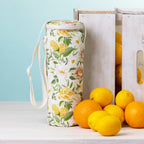 A floral bottle bag with a white strap stands on a white tabletop beside white wooden crates and bright lemons and oranges against a pale blue background.